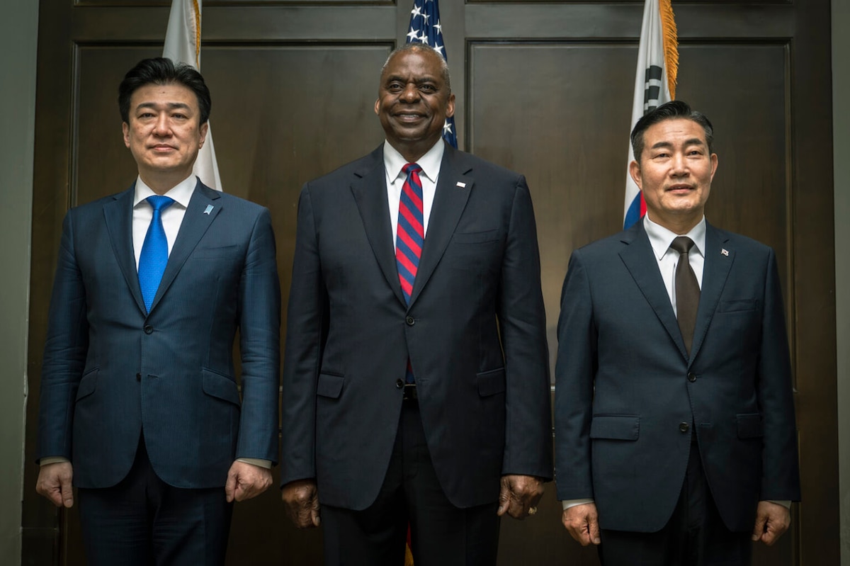 Three people wearing business suits stand in front of U.S., Japanese and South Korean flags.