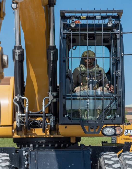 A U.S. Airman operates a construction vehicle, enclosed in a secure, barred cab, wearing protective gear and a helmet.