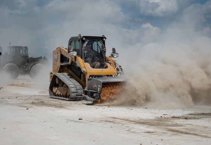 A U.S. Airman maneuvers a loader vehicle equipped with a sweeping attachment, kicking up dust as it cleans a large area.