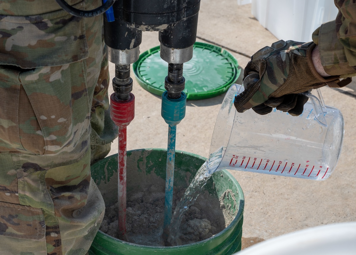 A close-up views of a U.S. Airman pouring water from a measuring jug into a bucket containing concrete mix.