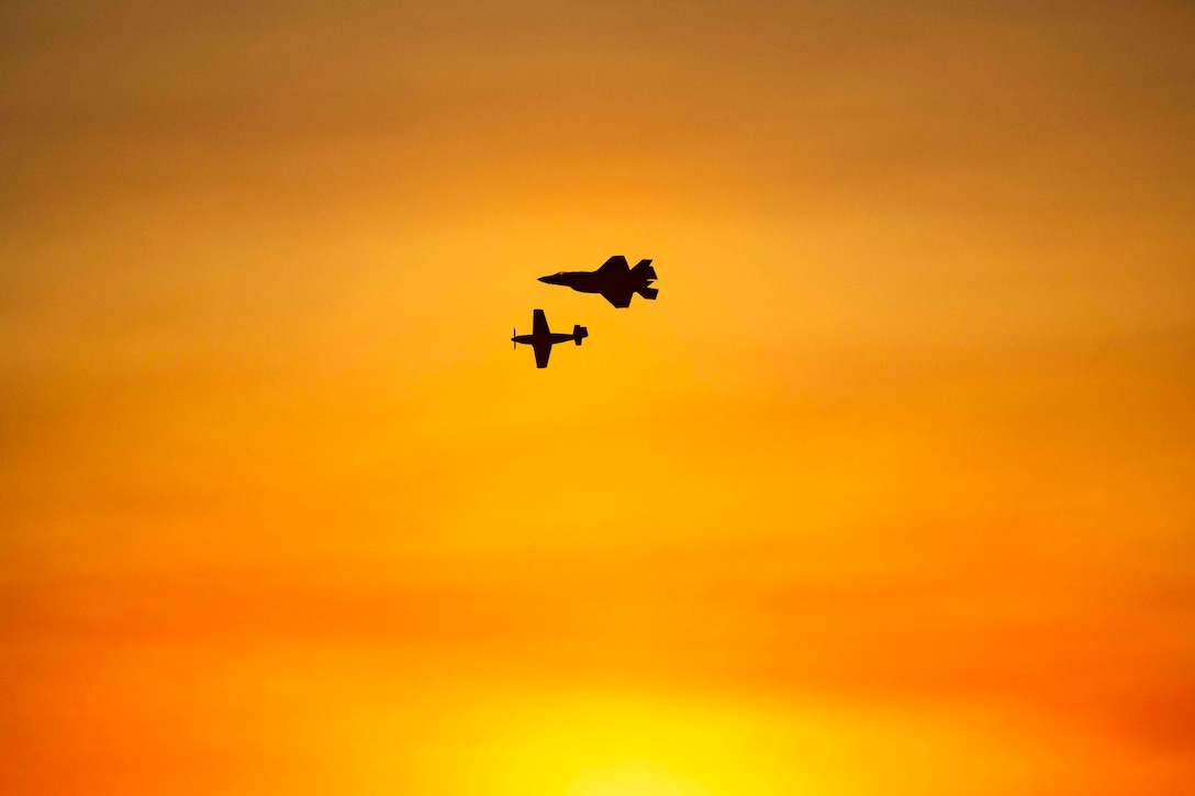 Two aircraft are silhouetted as they fly in formation across an orange sky.