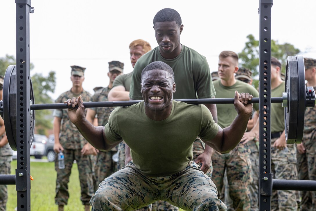 A Marine sweats and grimaces while squatting and lifting a heavy barbell as others stand and watch.