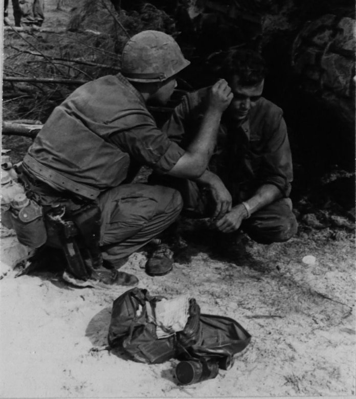 A man squats down to apply something to the side of another seated man’s head.