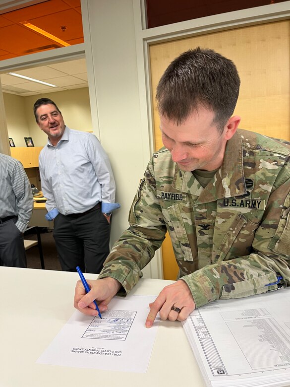 A man in an Army uniform signs a piece of paper while a man in black pants and a blue shirt watches in the background.