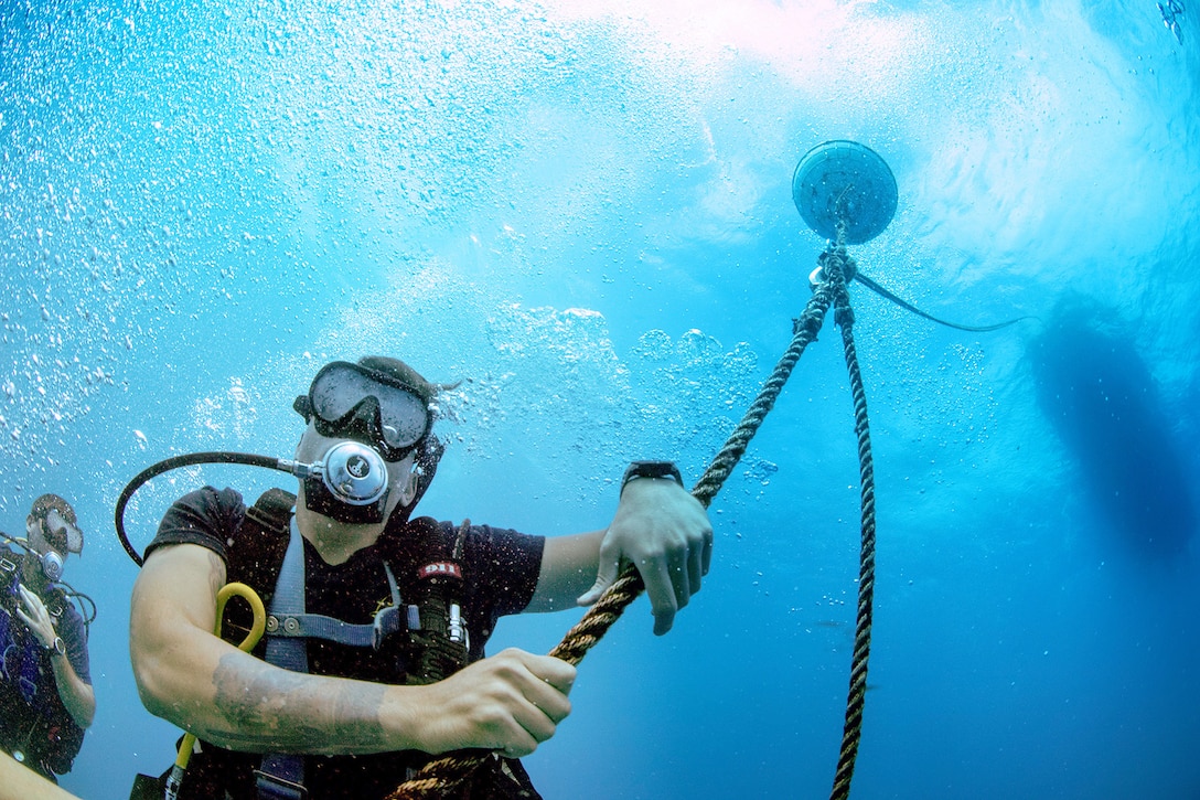 A sailor holds onto a rope underwater as another person swims in the background.