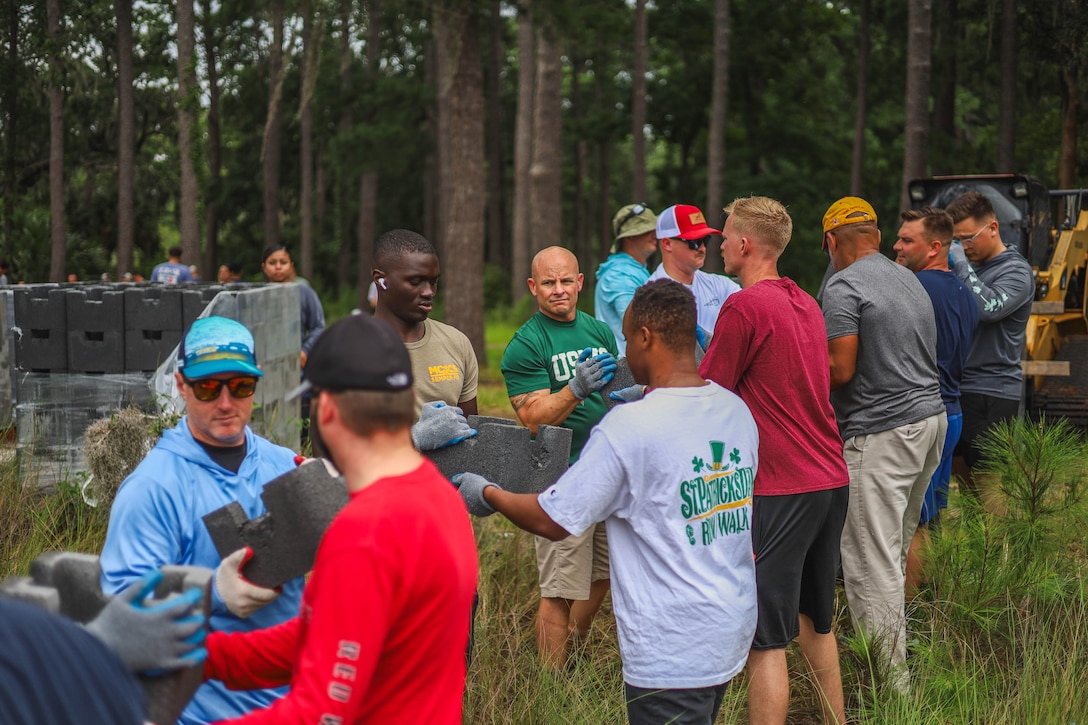 People in civilian attire face one another in parallel rows in a grassy area to pass along materials for building an oyster reef.