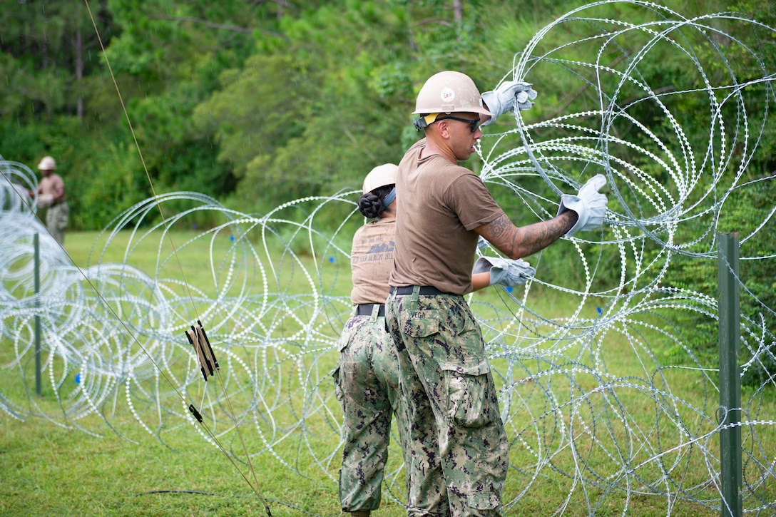 Two sailors wearing protective gloves and helmets set up barbed wire in a field.