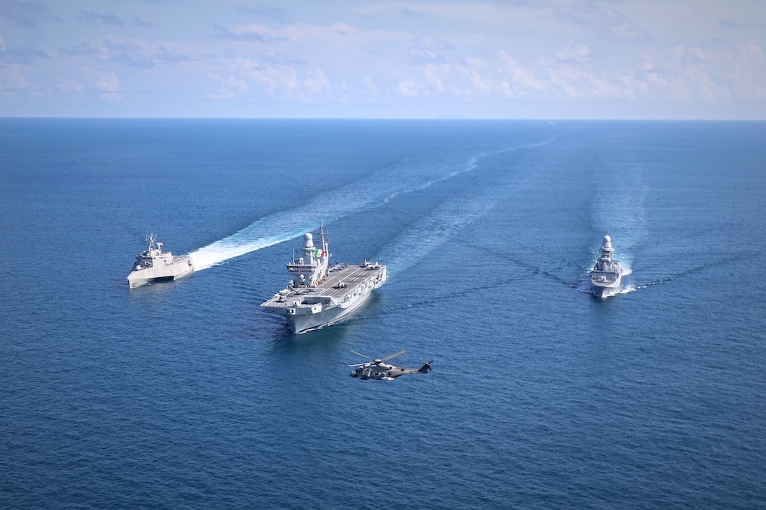 A helicopter flies in front of three ships sailing in formation in a body of water during daylight.