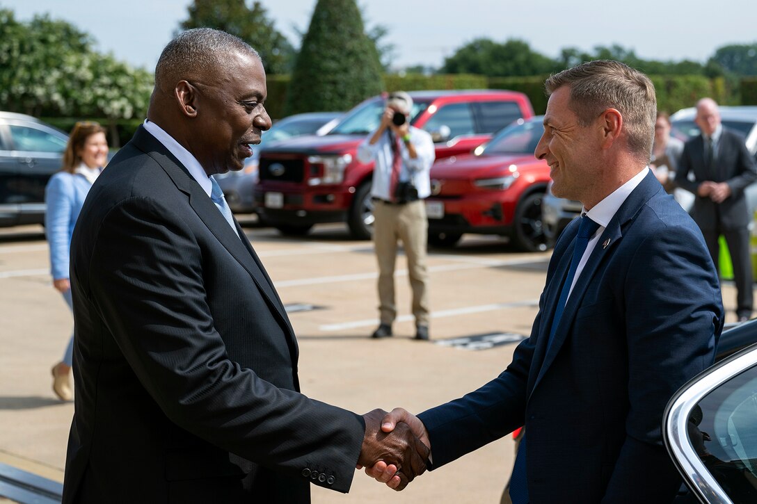 Two civilians shake hands in a parking lot in front of a photographer and other attendees.
