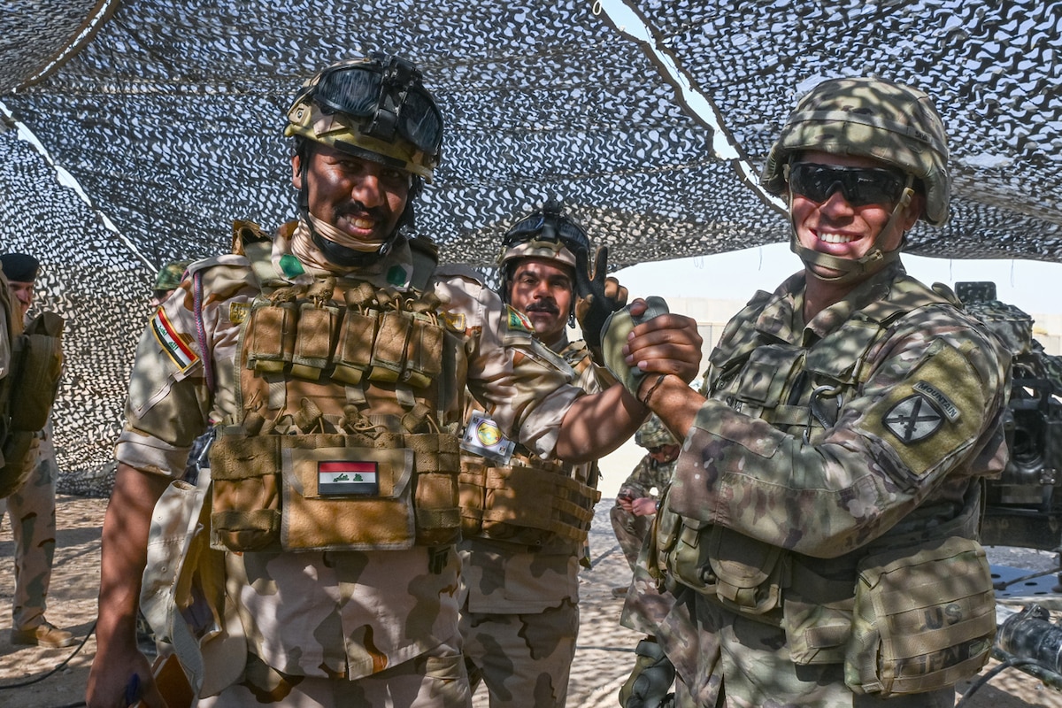 A U.S. and foreign service member wearing battle gear shake hands under camouflage netting.