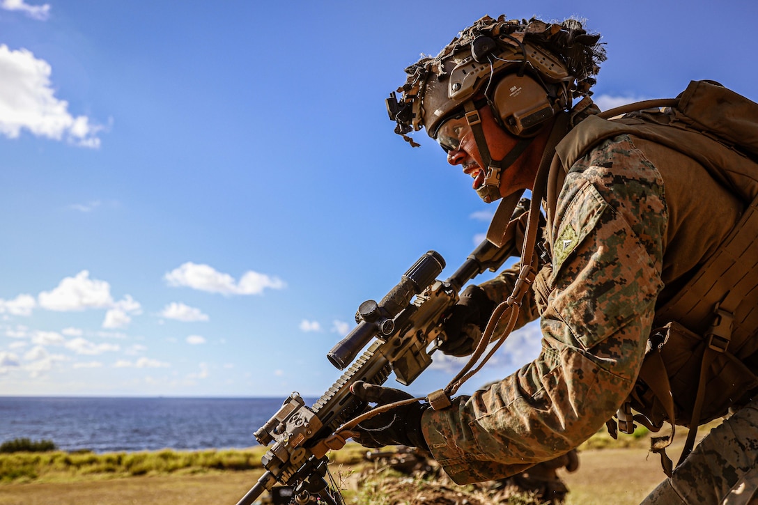 A Marine holding a weapon looks forward while training in a large field near the ocean.