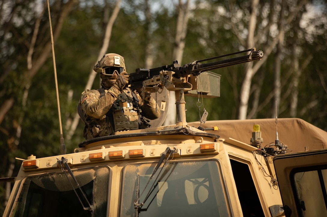 A soldier wearing dark goggles stands in the cab of a military vehicle holding a machine gun.