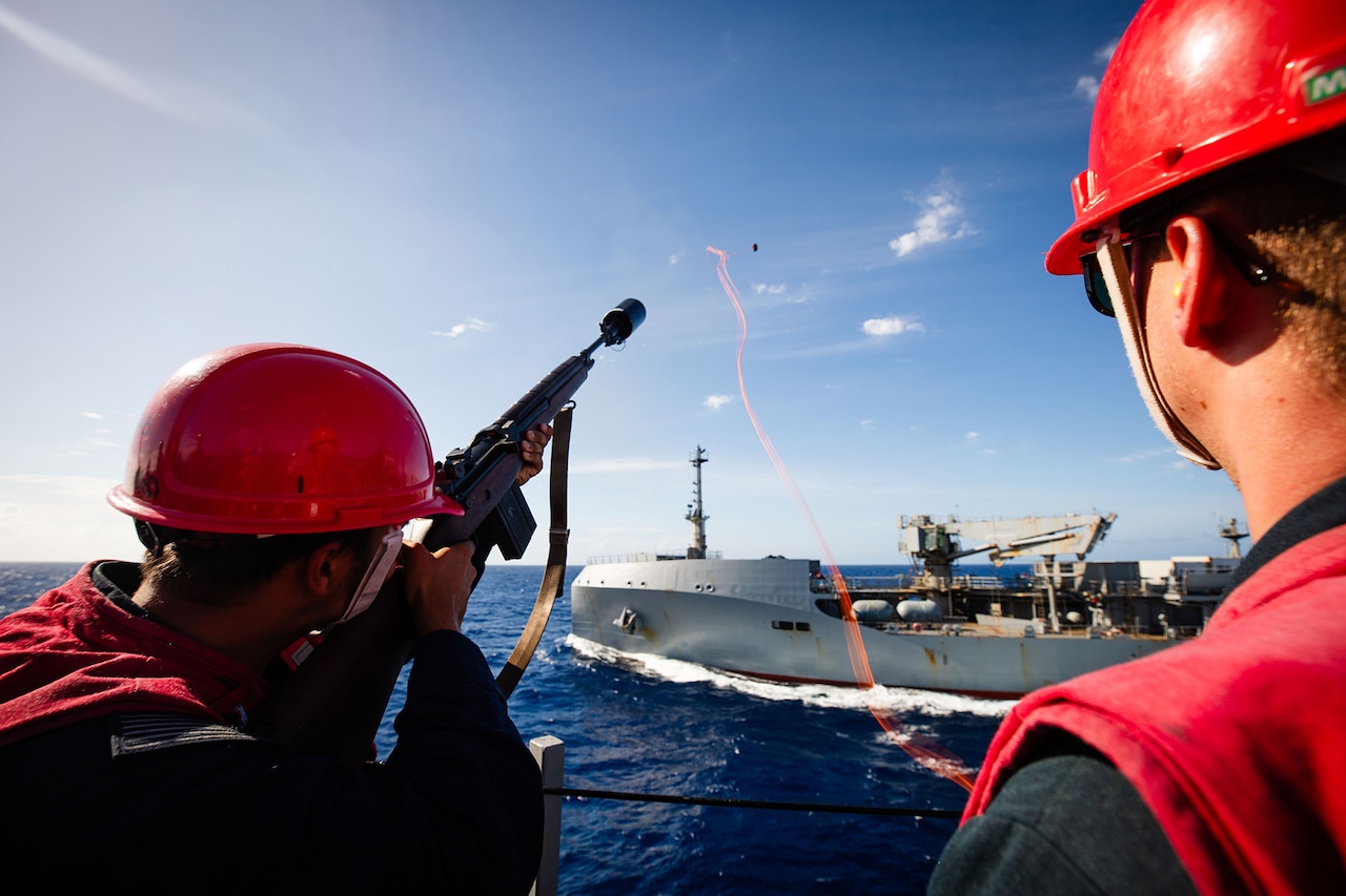 A sailor fires a shot line into the sky at another military ship at sea while another sailor looks on.