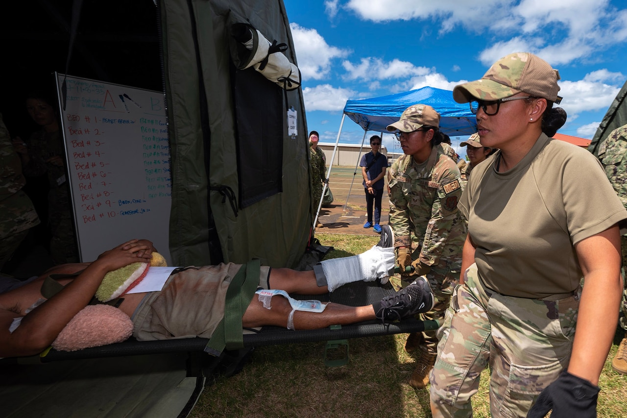 Two civilians in military uniform hold a stretcher with a simulated patient halfway into a tent.
