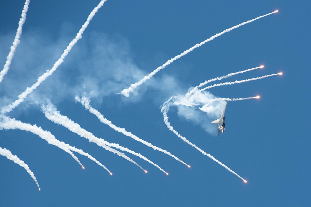 A military aircraft performs a maneuver against a bright blue sky, creating vapor and contrails.