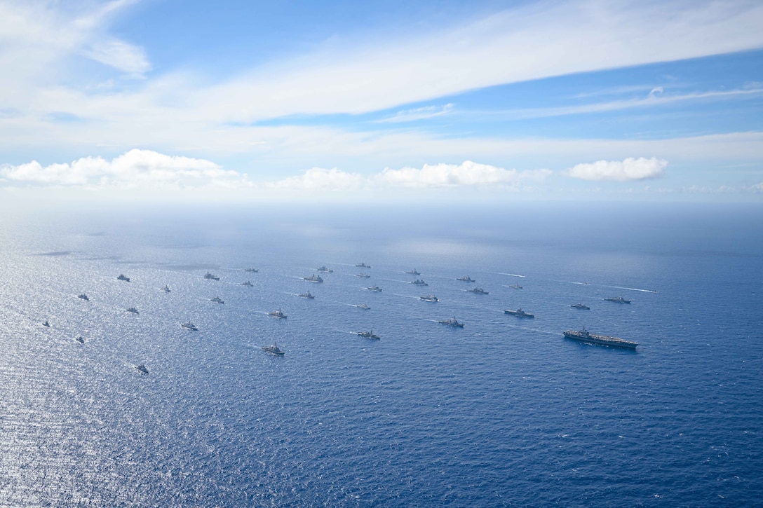 A ship leads dozens of other ships as they sail in formation in a body of water under a blue sky.