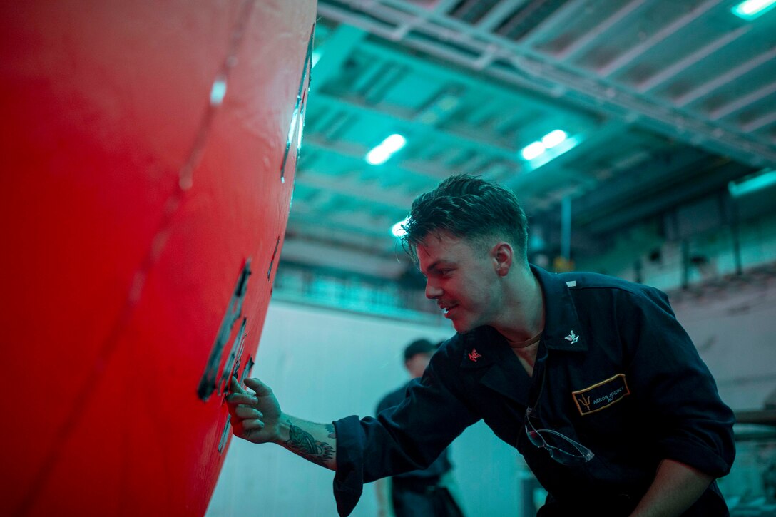 A sailor works on an orange inflatable device in a dimly lit garage-like area aboard a ship.