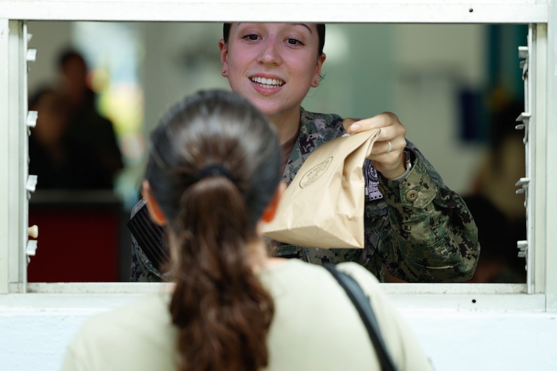 A sailor hands a paper bag to a patient through the open window of a structure.