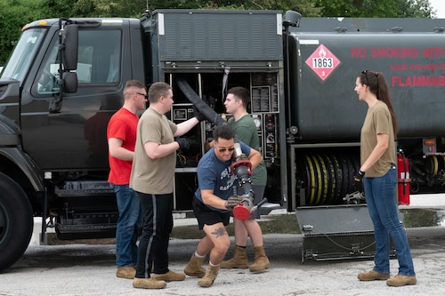 An Airman begins to drag a hose away from its holster as other Airman help uncurl it.