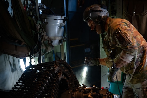 Airman with helmet shines flashlight through small cloud of smoke while inspecting large gun aboard plane