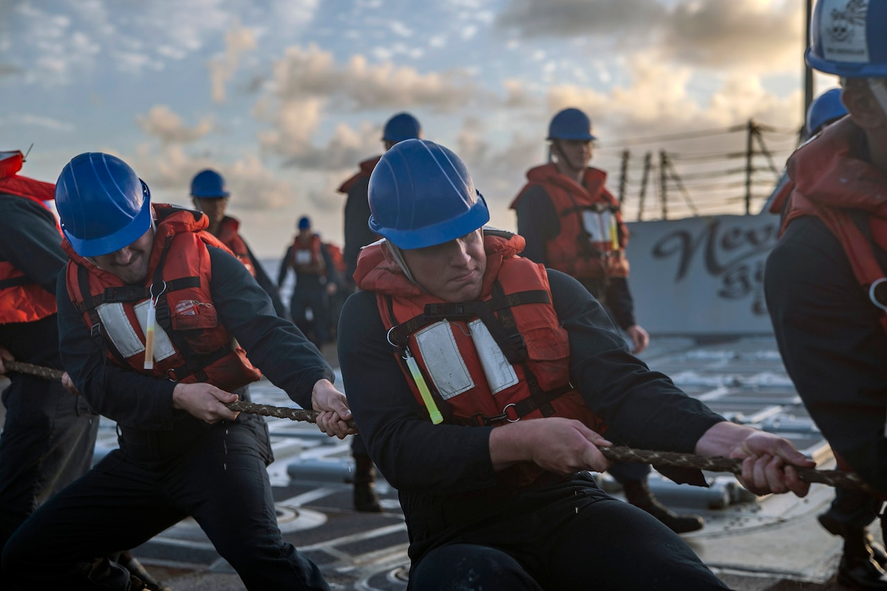 A group of sailors wearing life vests on the deck of a warship heave a line while other participants mill about in the backgound.