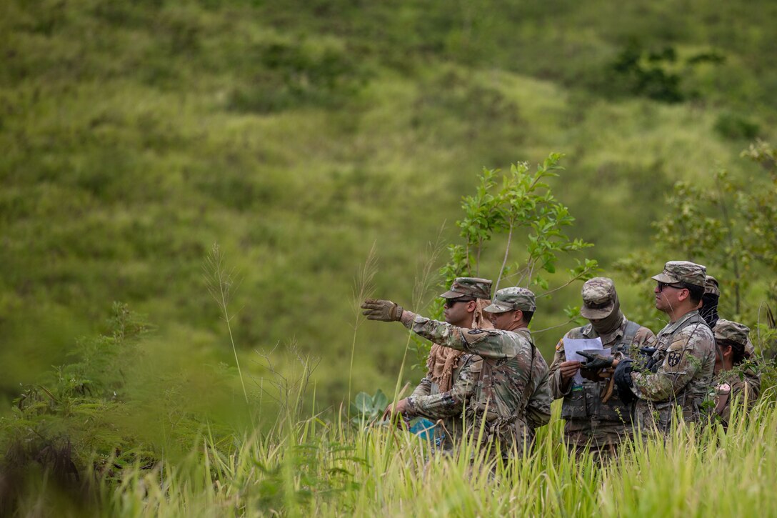 A group of soldiers stand in an overgrown field as one points forward and another reads a sheet of paper.