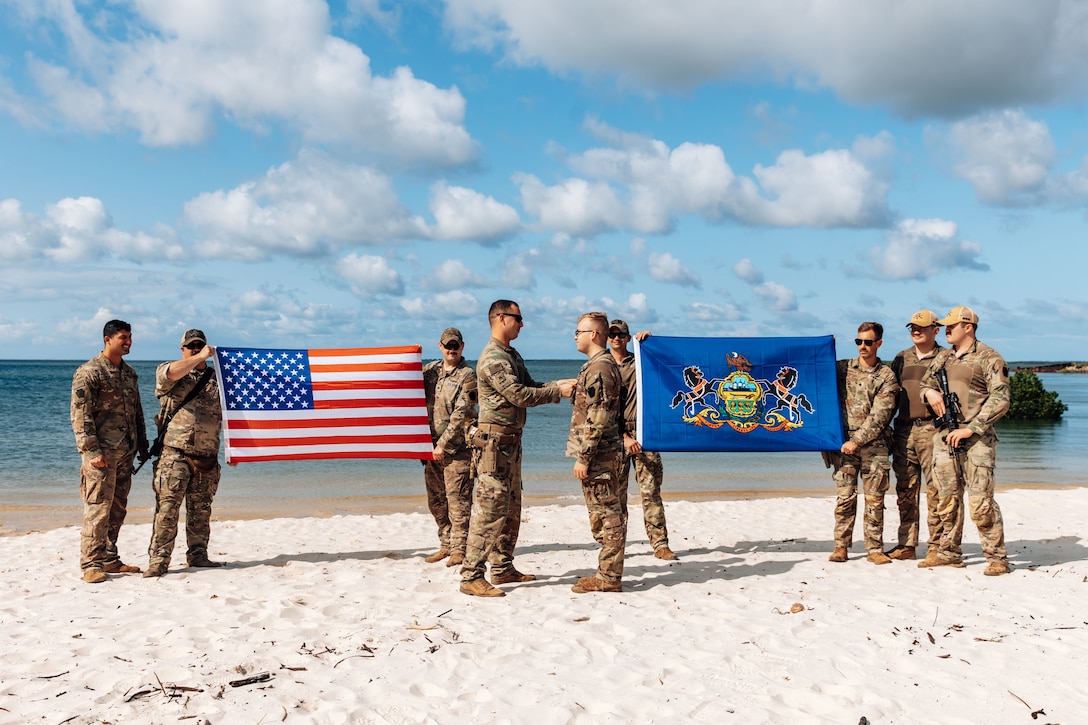 Two soldiers face each other during a ceremony on a beach while other soldiers hold two large flags and watch.