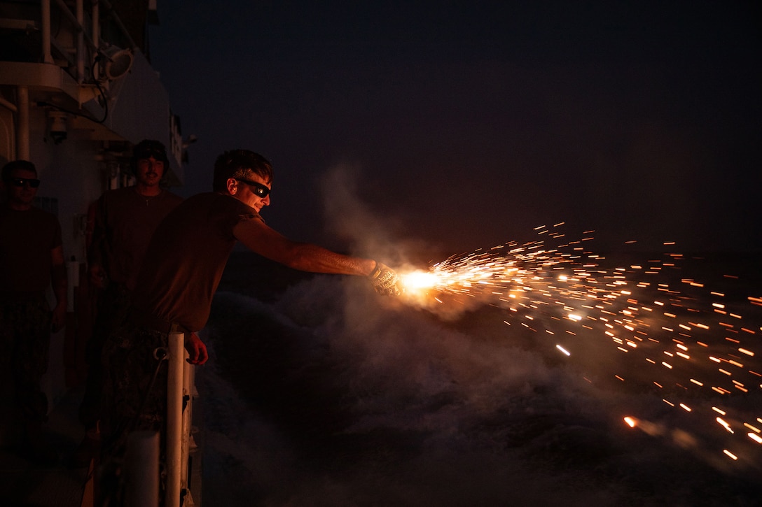 A service member wearing protective glasses holds a flare emitting yellow sparks from the side of a ship at night.