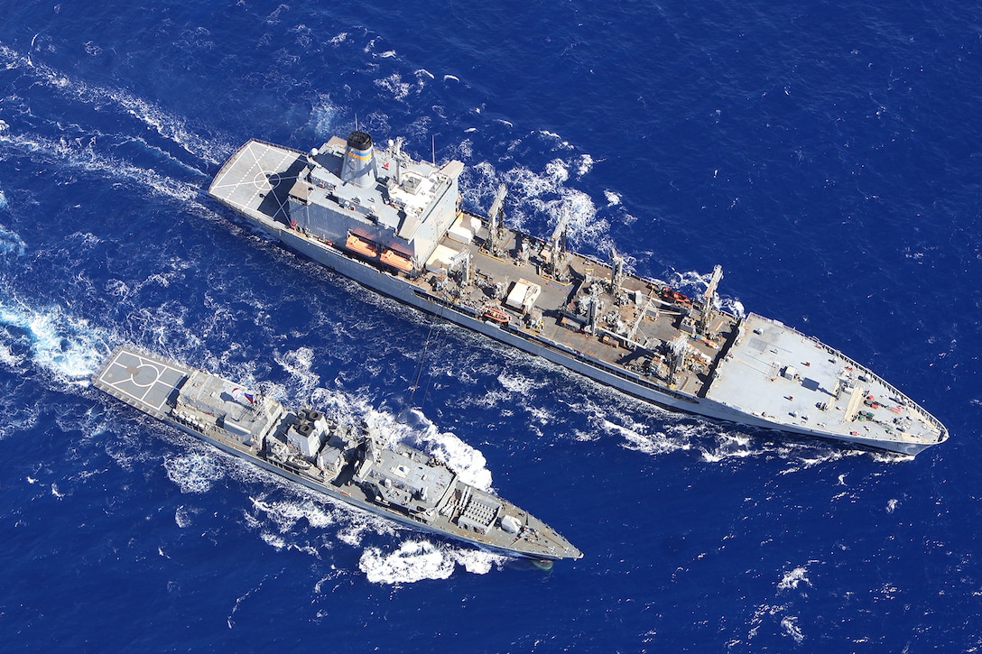 A smaller vessel sails alongside a larger ship in open water during daylight creating a wake behind them.