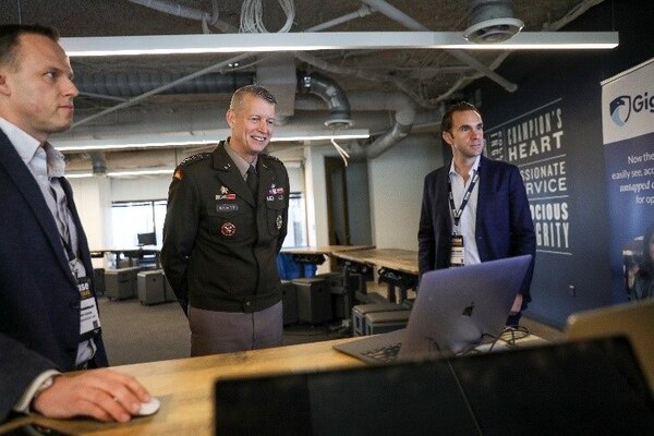 A service member in uniform and two people in civilian attire stand near a computer.