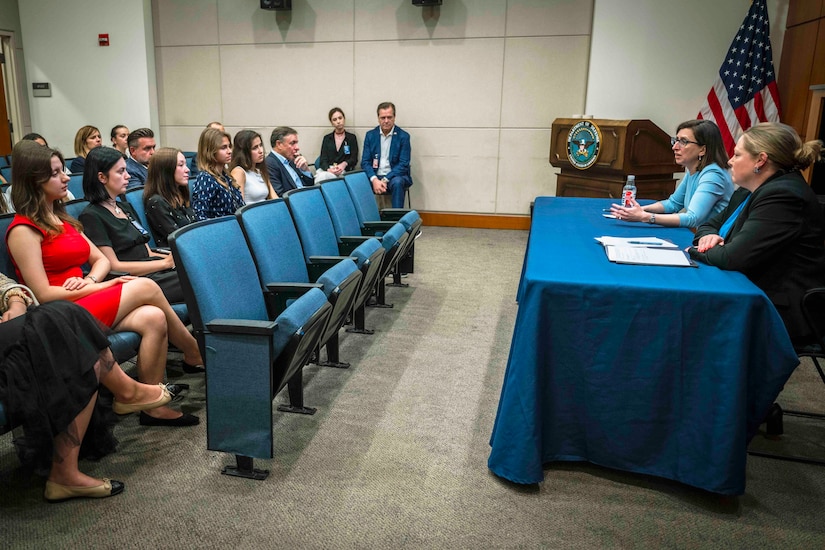 Two individuals sit at a table in front of a room. In front of them, other individuals are seated in chairs.