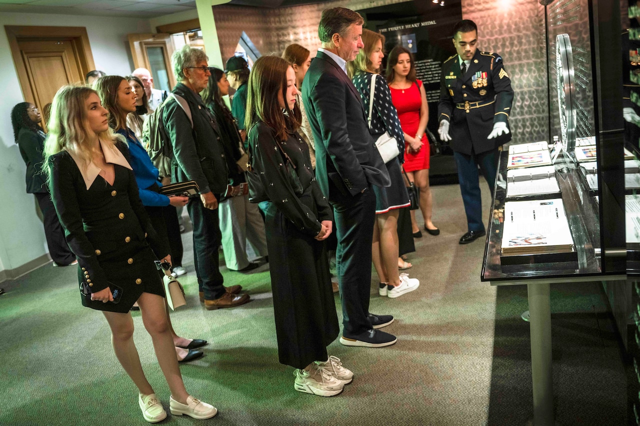 More than a dozen individuals stand in a small, museum-like room and look at artifacts.