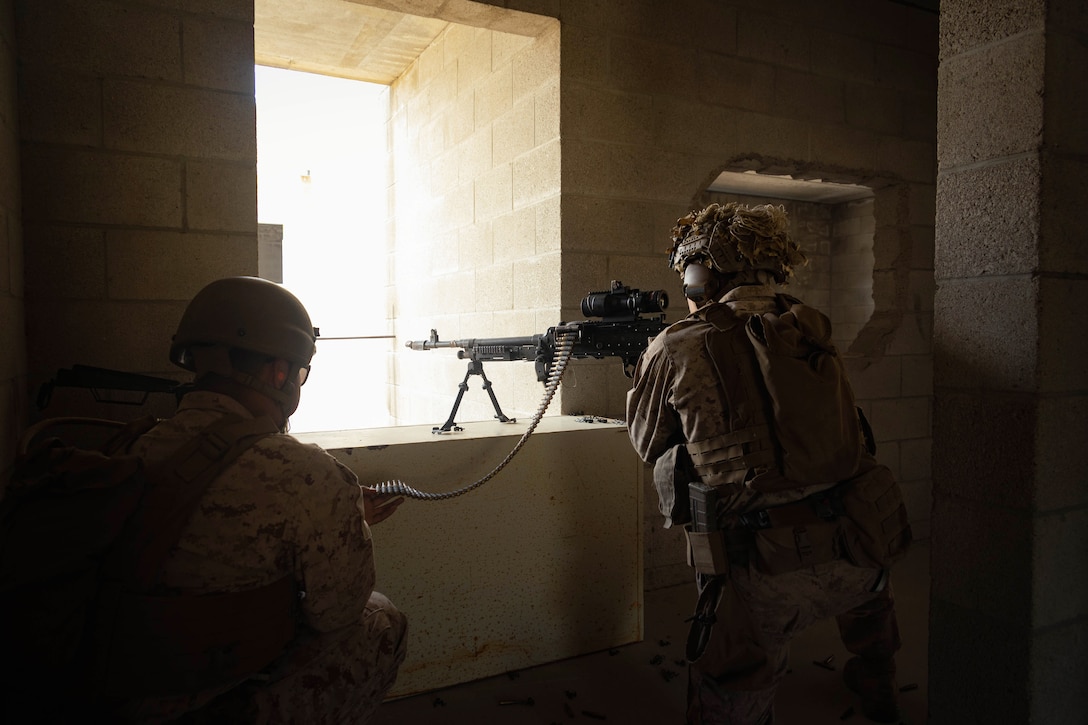 A Marine looks through the scope of a weapon positioned in front of a window displaying bright light as a fellow Marine kneels to the left holding ammunition.