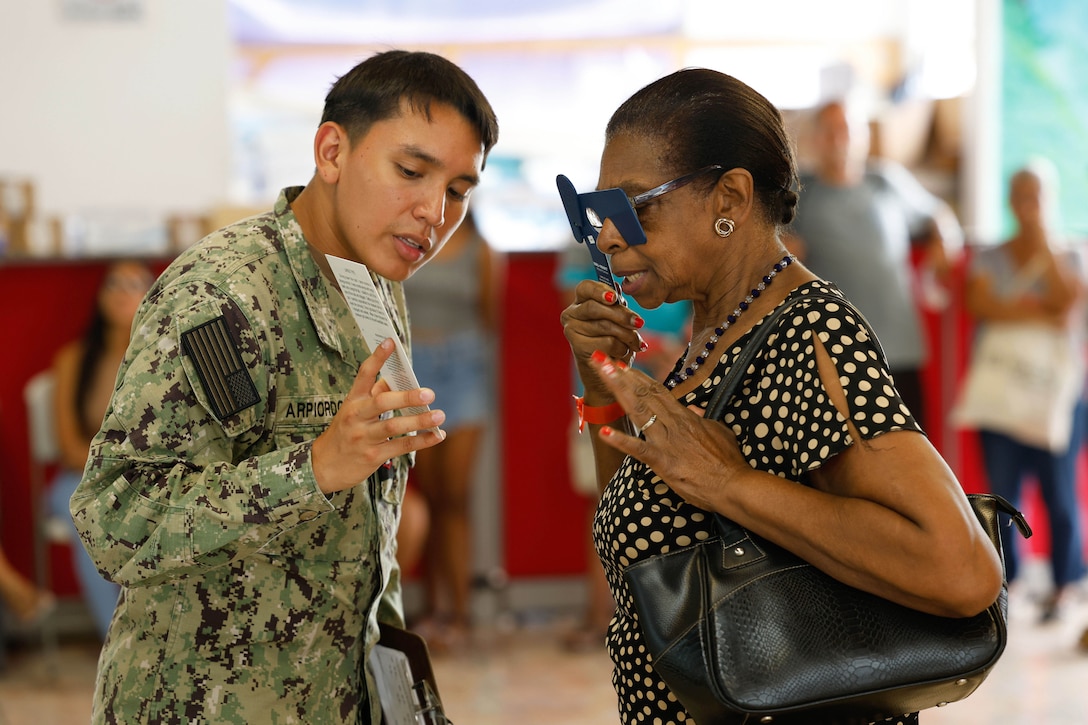 A patient uses a device to cover one of their eyes while reading from a card in a sailor’s hand as civilians stand in formation in a blurred background.