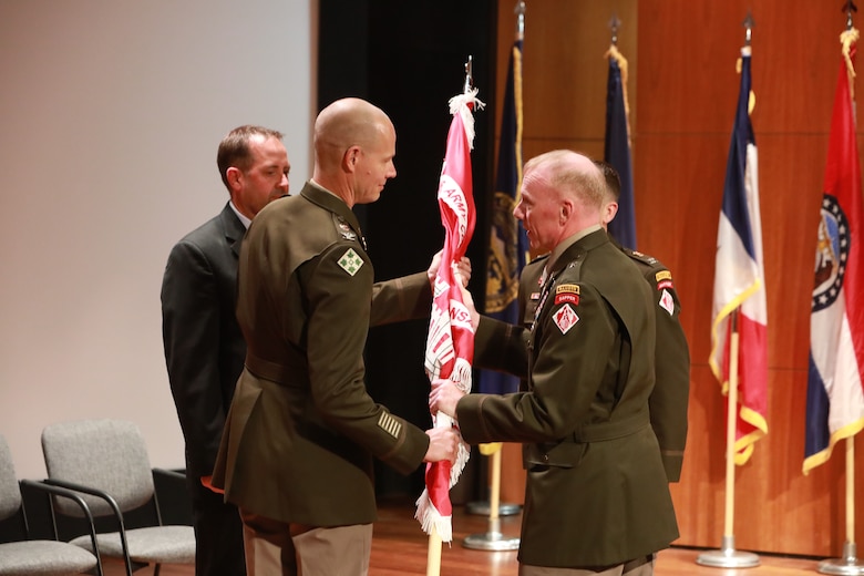 Three men in Army dress uniforms stand with a man in a black suit. One man in Army dress uniform passes a red flag to another man in Army dress uniform.