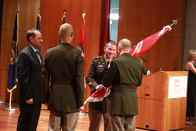 Three men in Army dress uniforms stand with a man in a black suit. One man in Army dress uniform passes a red flag to another man in Army dress uniform.