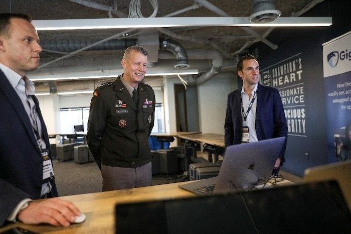 A service member in uniform and two people in civilian attire stand near a computer.