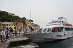 Tourists depart boat via ramp, with buildings and trees on a hill in the background.
