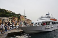 Tourists depart boat via ramp, with buildings and trees on a hill in the background.