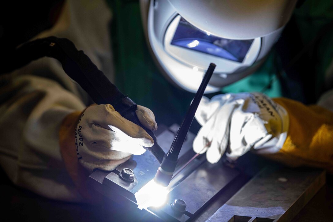 A close-up  photo shows an airman wearing protective headgear, clothing and gloves uses a welding tool on a metal object. A small flashlight lights up the area.