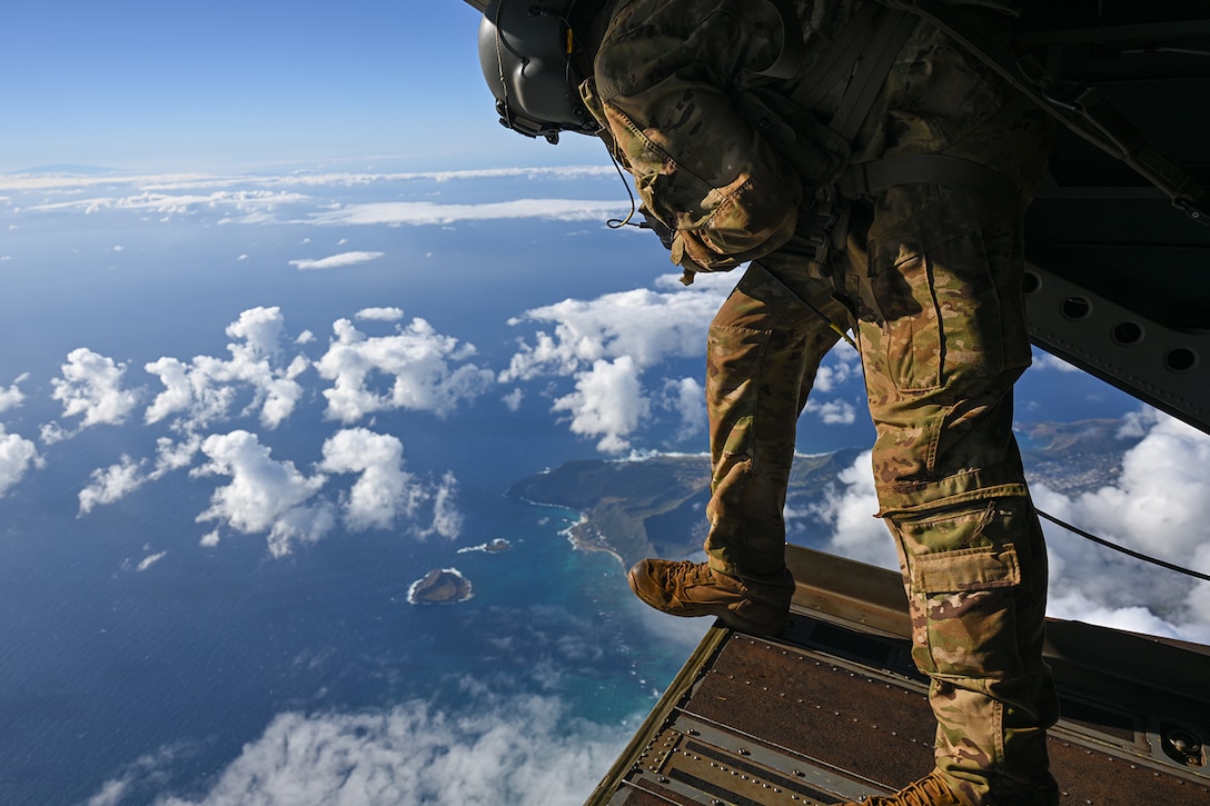 A soldier looks out of the opening of a military aircraft while in flight during daylight over a body of water. Clouds can be seen beneath the aircraft.