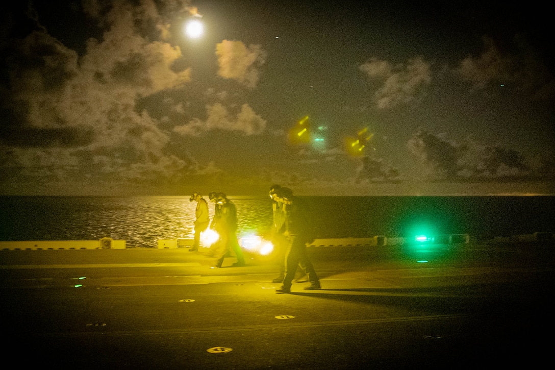 A group of sailors walk along the flight deck of a Navy ship at night while using flashlights emitting a yellow glow. The deck is illuminated by a green light.