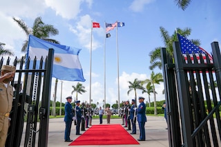 Military personnel salute in front of flags at the end of a red carpet flanked by a military ceremonial honor guard personnel.