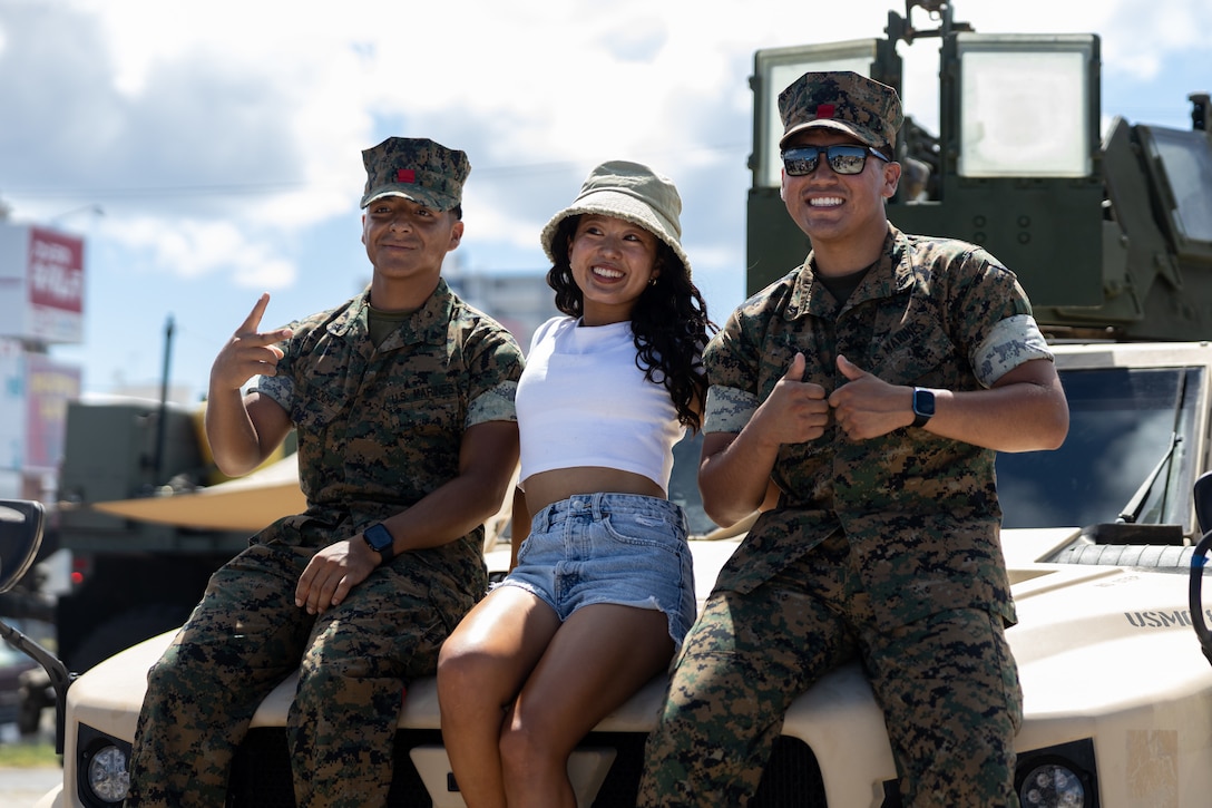 U.S. Marine Corps Lance Cpl. Michel Morocho, left, and Lance Cpl. Takenya Yazzie, both motor transport operators with 3rd Landing Support Battalion, 3rd Marine Logistics Group, pose for a group photo with a festival attendee on a Joint Light Tactical Vehicle during the Camp Foster Festival on Camp Foster, Okinawa, Japan, July 6, 2024. Marine Corps Community Services Okinawa hosted this open gate event to bring local residents and Status of Forces Agreement members together during the Fourth of July weekend. The festival featured live entertainment, food trucks, and a multitude of activities. Morocho is a native of New York and Yazzie is a native of New Mexico. (U.S. Marine Corps photo by Lance Cpl. Skylia Waters-Hewitt)