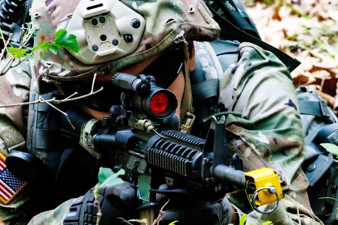 A soldier looks through the scope of a weapon while laying in the woods.