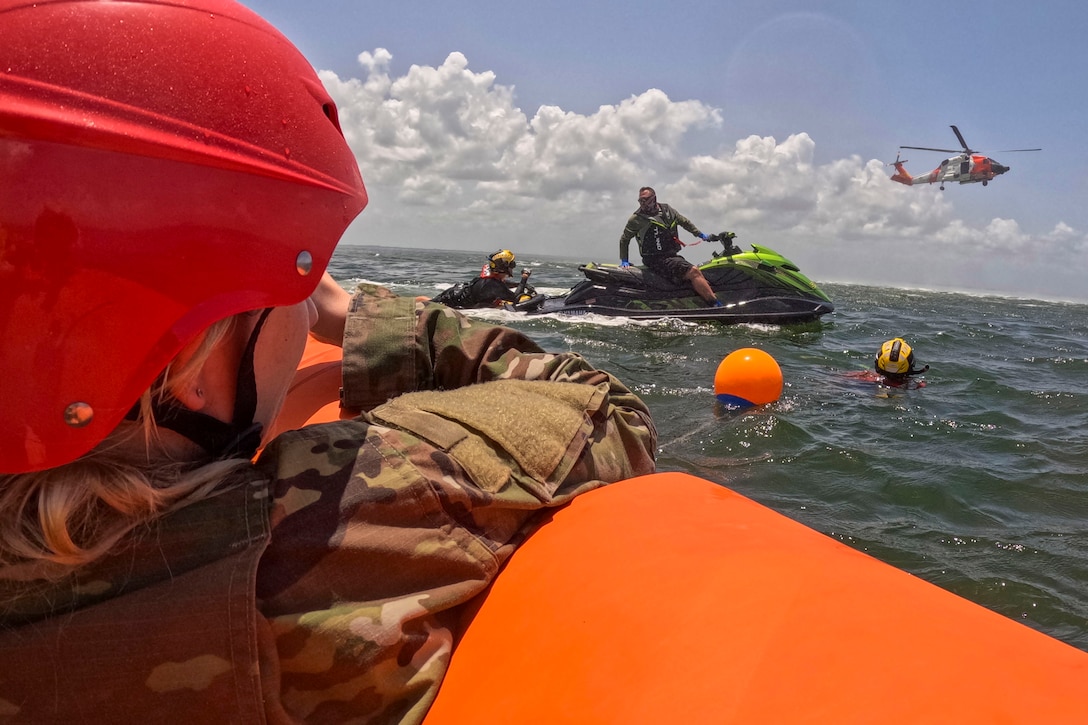 An airman wearing a red helmet looks out of a partially visible orange boat as a fellow airman floats in a body of water near a jet ski and hovering helicopter.