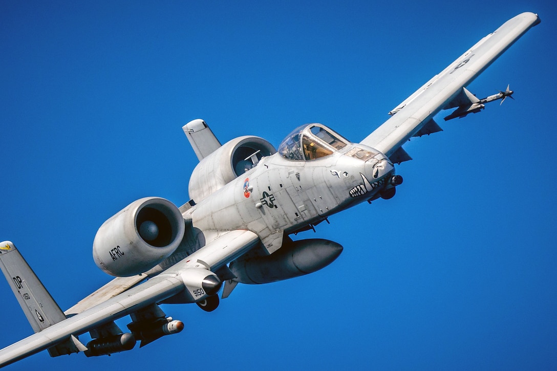 A close-up shows a military aircraft flying through a blue sky.