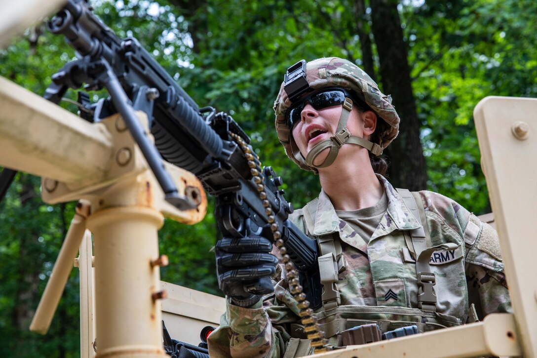 A soldier wearing dark glasses and gloves stands in the cab of a military vehicle and holds a machine gun.