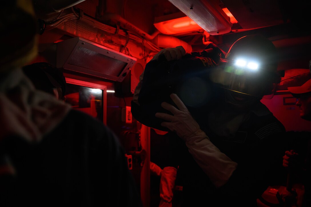 A sailor wearing a headlamp and helmet carries an object in a dark room illuminated by red light.