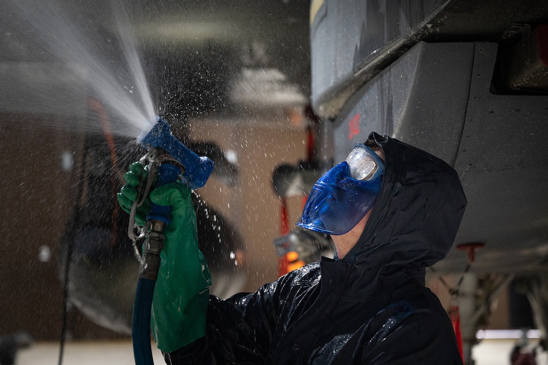 An airman wearing goggles and protective gear uses a hose to wash a military aircraft, creating a mist of water droplets.